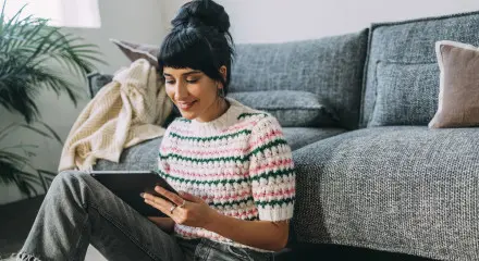 A woman sitting comfortably on a couch at home, smiling while using a tablet or device.