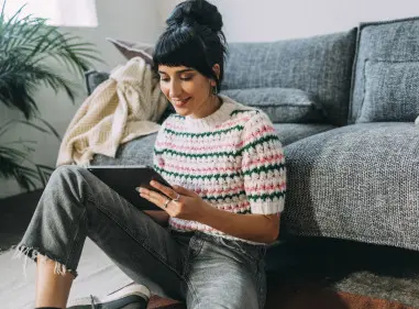 A woman sitting comfortably on a couch at home, smiling while using a tablet or device.