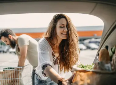 Young female packing reusable grocery bags into the boot. Male reaching for more groceries from a trolley in the background