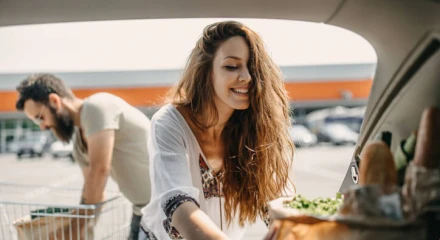 Young female packing reusable grocery bags into the boot. Male reaching for more groceries from a trolley in the background