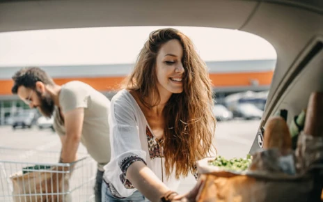 Young female packing reusable grocery bags into the boot. Male reaching for more groceries from a trolley in the background