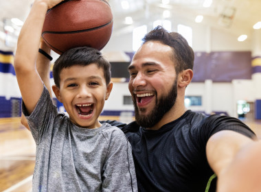 Young smiling boy holding a basketball on his head with smiling dad next to him taking a selfie of them both.