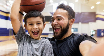 Young smiling boy holding a basketball on his head with smiling dad next to him taking a selfie of them both.