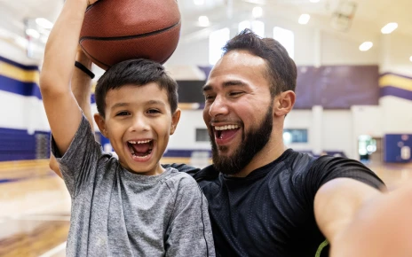 Young smiling boy holding a basketball on his head with smiling dad next to him taking a selfie of them both.