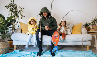 Young man and two kids dressed up in wet weather gear, sitting on the couch fishing for paper fish