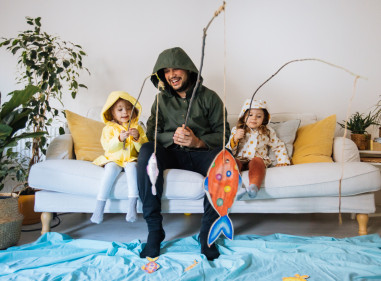 Young man and two kids dressed up in wet weather gear, sitting on the couch fishing for paper fish