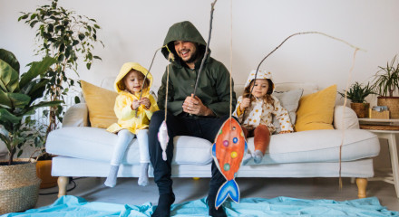 Young man and two kids dressed up in wet weather gear, sitting on the couch fishing for paper fish