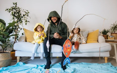 Young man and two kids dressed up in wet weather gear, sitting on the couch fishing for paper fish