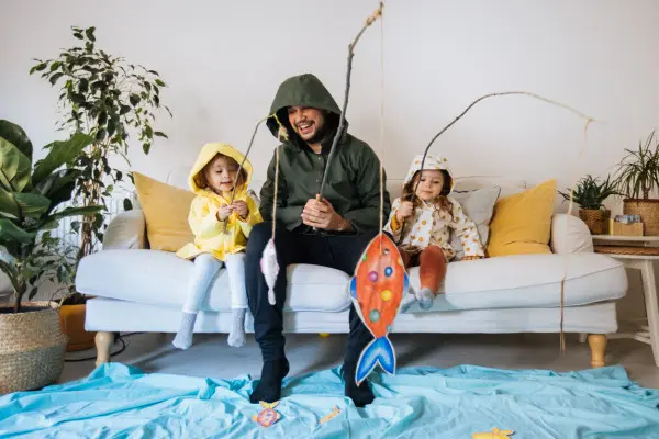 Young man and two kids dressed up in wet weather gear, sitting on the couch fishing for paper fish