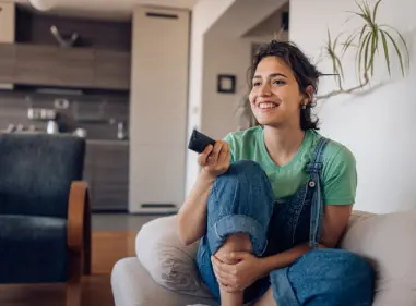 Smiling young woman sitting on her couch clicking the remote to change the channel