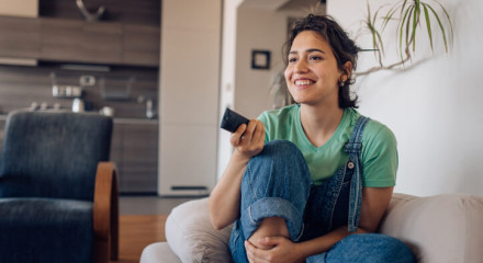Smiling young woman sitting on her couch clicking the remote to change the channel