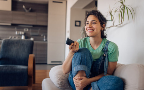 Smiling young woman sitting on her couch clicking the remote to change the channel