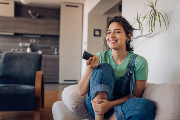 Smiling young woman sitting on her couch clicking the remote to change the channel