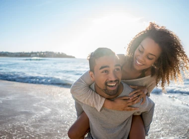 Smiling young man piggy-backing smiling partner in shallow waves at the beach