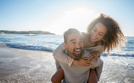 Smiling young man piggy-backing smiling partner in shallow waves at the beach