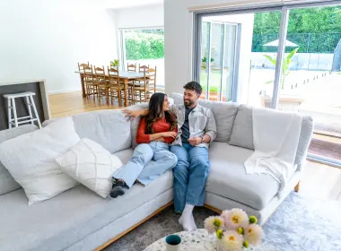 Two people sitting on a sofa in a bright, modern open-plan living and dining area with large windows.