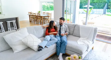 Two people sitting on a sofa in a bright, modern open-plan living and dining area with large windows.