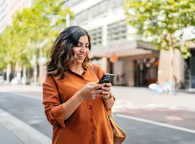 Woman using a smartphone on a tree-lined city street with buildings in the background.