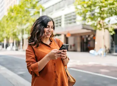 Woman using a smartphone on a tree-lined city street with buildings in the background.