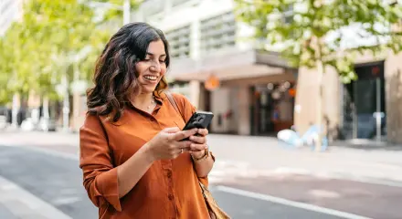 Woman using a smartphone on a tree-lined city street with buildings in the background.