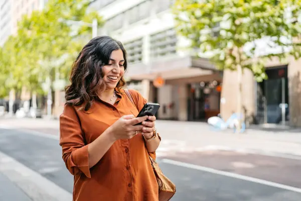 Woman using a smartphone on a tree-lined city street with buildings in the background.