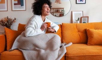 Woman sitting on couch with a blanket and holding a mug laughing