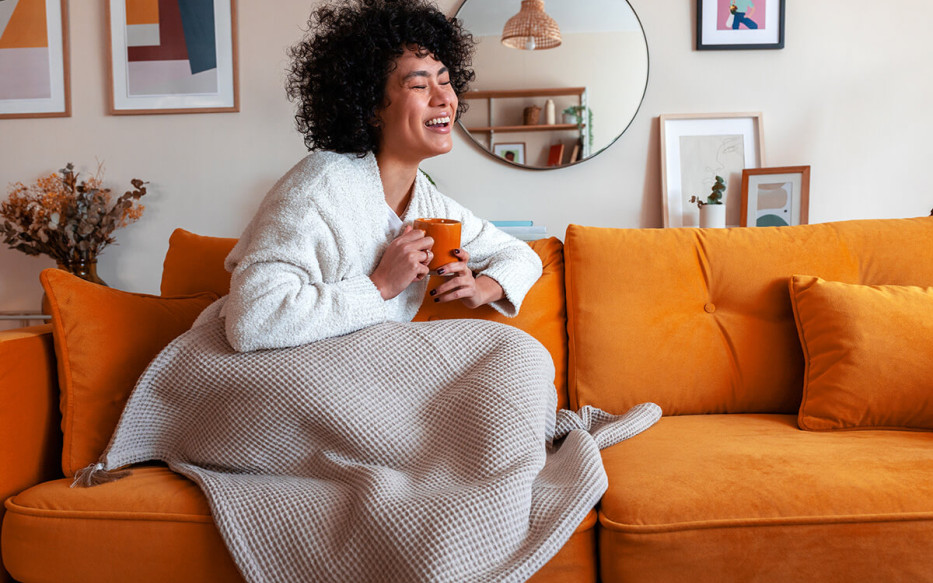 Woman sitting on couch with a blanket and holding a mug laughing