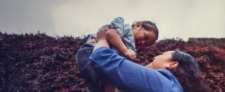 A person in a blue shirt joyfully lifts a smiling child in a denim outfit against a cloudy sky backdrop, conveying warmth and happiness.