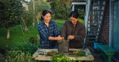 Two women smiling outdoors in a garden sorting compost into a compost bin.
