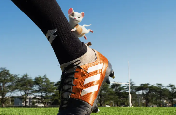 Illustration of Sorted's mouse holding onto the back of a rugbier's leg wearing black socks and orange sports shoes.