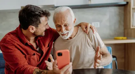 A younger adult sitting beside an older adult at a table, showing a smartphone, with an arm around the older adult’s shoulder in a home setting.