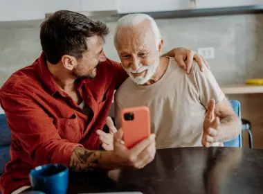 A younger adult sitting beside an older adult at a table, showing a smartphone, with an arm around the older adult’s shoulder in a home setting.