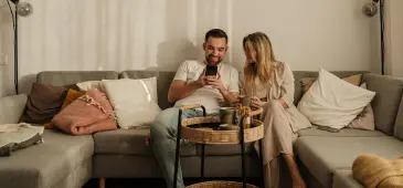 Two people sitting side by side on a grey sectional sofa in a softly lit living room, looking at a smartphone held by one person.