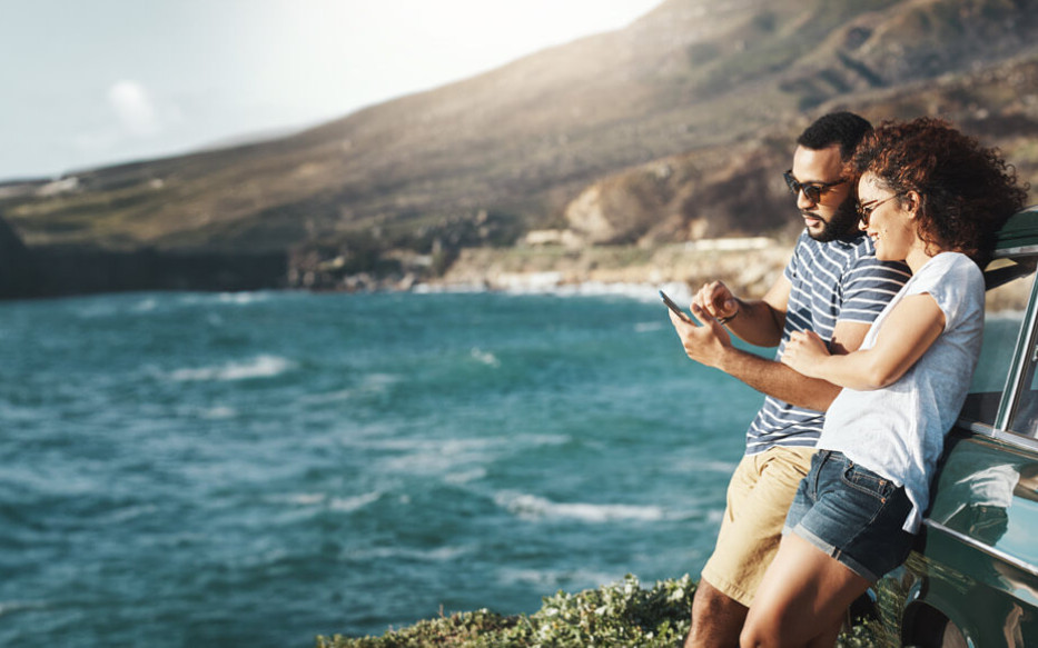 Two people are leaning against a car looking at their phone with the coastline in their background.