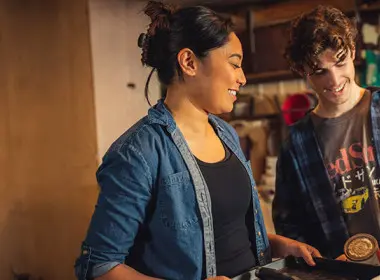 A man and woman are standing together in a garage holding up painting supplies and smiling
