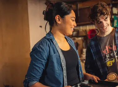 A man and woman are standing together in a garage holding up painting supplies and smiling