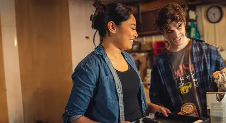A man and woman are standing together in a garage holding up painting supplies and smiling