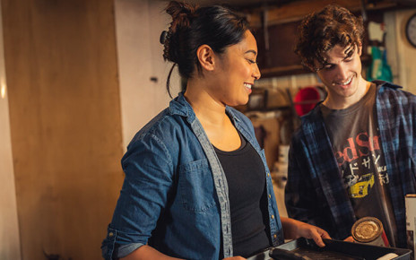 A man and woman are standing together in a garage holding up painting supplies and smiling