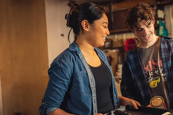 A man and woman are standing together in a garage holding up painting supplies and smiling