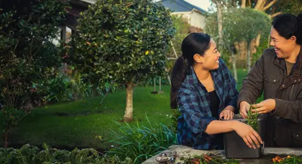 Two women are standing together outside and gardening. They are looking at each other and laughing.