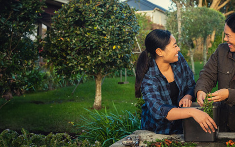 Two women are standing together outside and gardening. They are looking at each other and laughing.