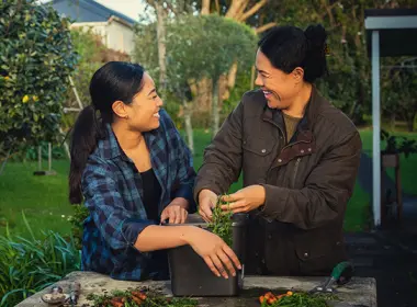 Two women are standing together outside and gardening. They are looking at each other and laughing.
