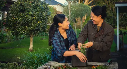 Two women are standing together outside and gardening. They are looking at each other and laughing.