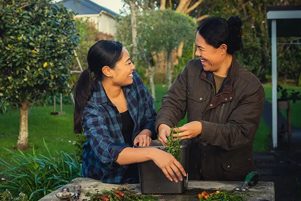 Two women are standing together outside and gardening. They are looking at each other and laughing.