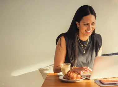 A woman is sitting at a cafe table typing on her laptop with a coffee and pastry to the side.
