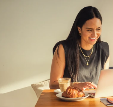 A woman is sitting at a cafe table typing on her laptop with a coffee and pastry to the side.