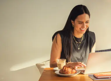 A woman is sitting at a cafe table typing on her laptop with a coffee and pastry to the side.