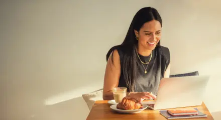 A woman is sitting at a cafe table typing on her laptop with a coffee and pastry to the side.