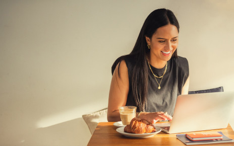 A woman is sitting at a cafe table typing on her laptop with a coffee and pastry to the side.
