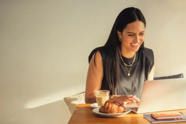 A woman is sitting at a cafe table typing on her laptop with a coffee and pastry to the side.